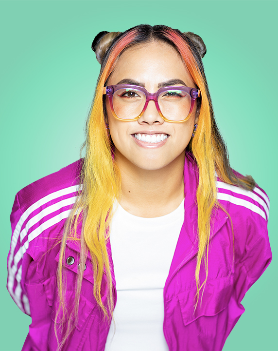 Persona con pelo amarillo y rosa, gafas a juego y una llamativa chaqueta rosa haciendo un guiño a la cámara