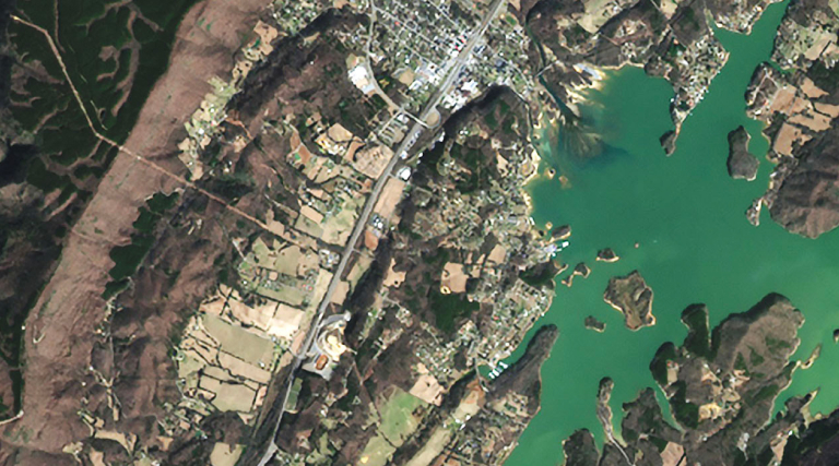 Imagen de satélite de una ciudad costera en la confluencia de un río con una masa de agua, rodeada de zonas verdes y terrenos talados