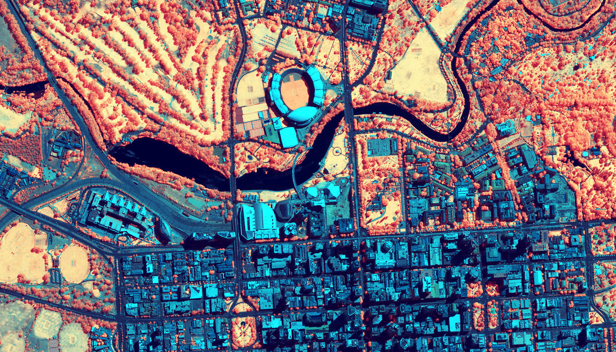 Mapa por satélite codificado por colores de una ciudad con tonos azules, naranjas y rojos, que muestra un río, espacios verdes y una estructura similar a un estadio en un trazado urbano en cuadrícula.