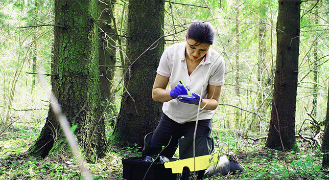 Una foto de una persona vestida de manera informal arrodillada en un bosque tomando notas en un dispositivo móvil con una pequeña variedad de equipamiento esparcido por el suelo