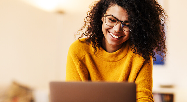 Una persona joven con gafas y jersey amarillo sonriendo mientras contempla el monitor de un ordenador portátil
