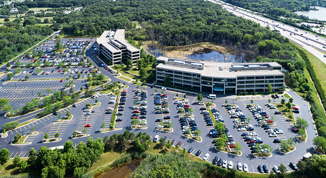 Una vista aérea de un edificio corporativo y un aparcamiento lleno de coches construidos cerca de un estanque y una zona boscosa con una autovía justo detrás